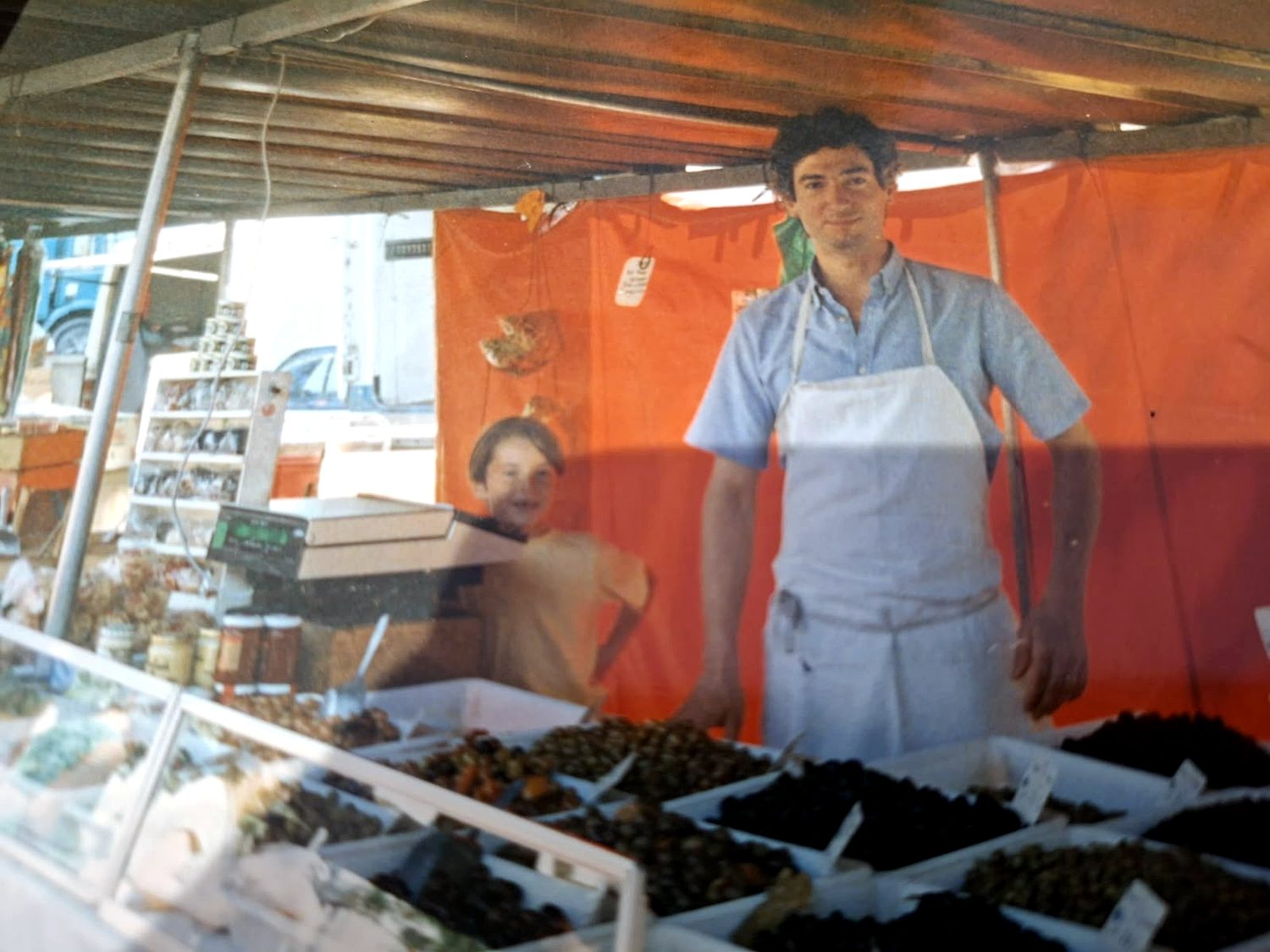 Olivier Daire à l’étal d’olives sous l’auvent orange, un enfant à ses côtés